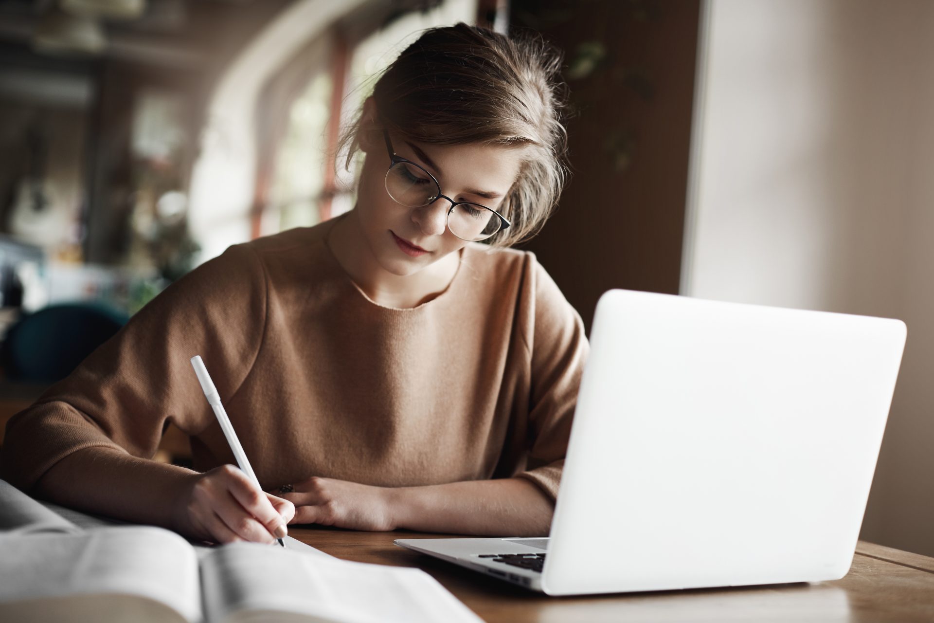 hardworking focused woman trendy glasses concentrating writing essay sitting cozy cafe near laptop working making notes carefully 36587